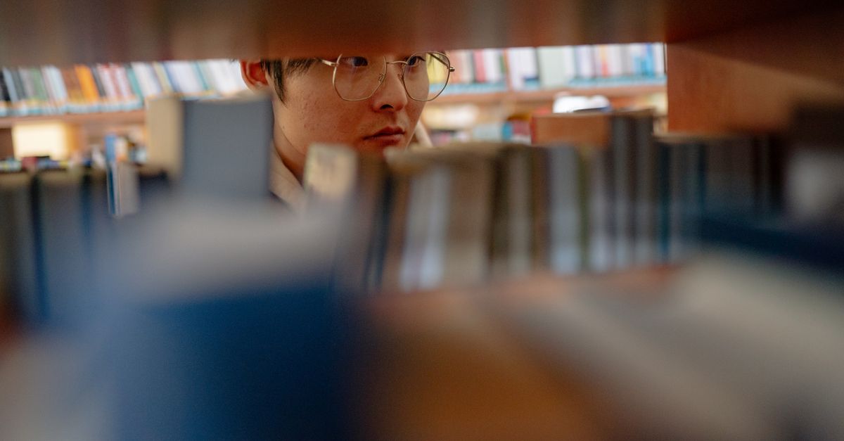 Young man wearing glasses studying intently between library shelves filled with books.